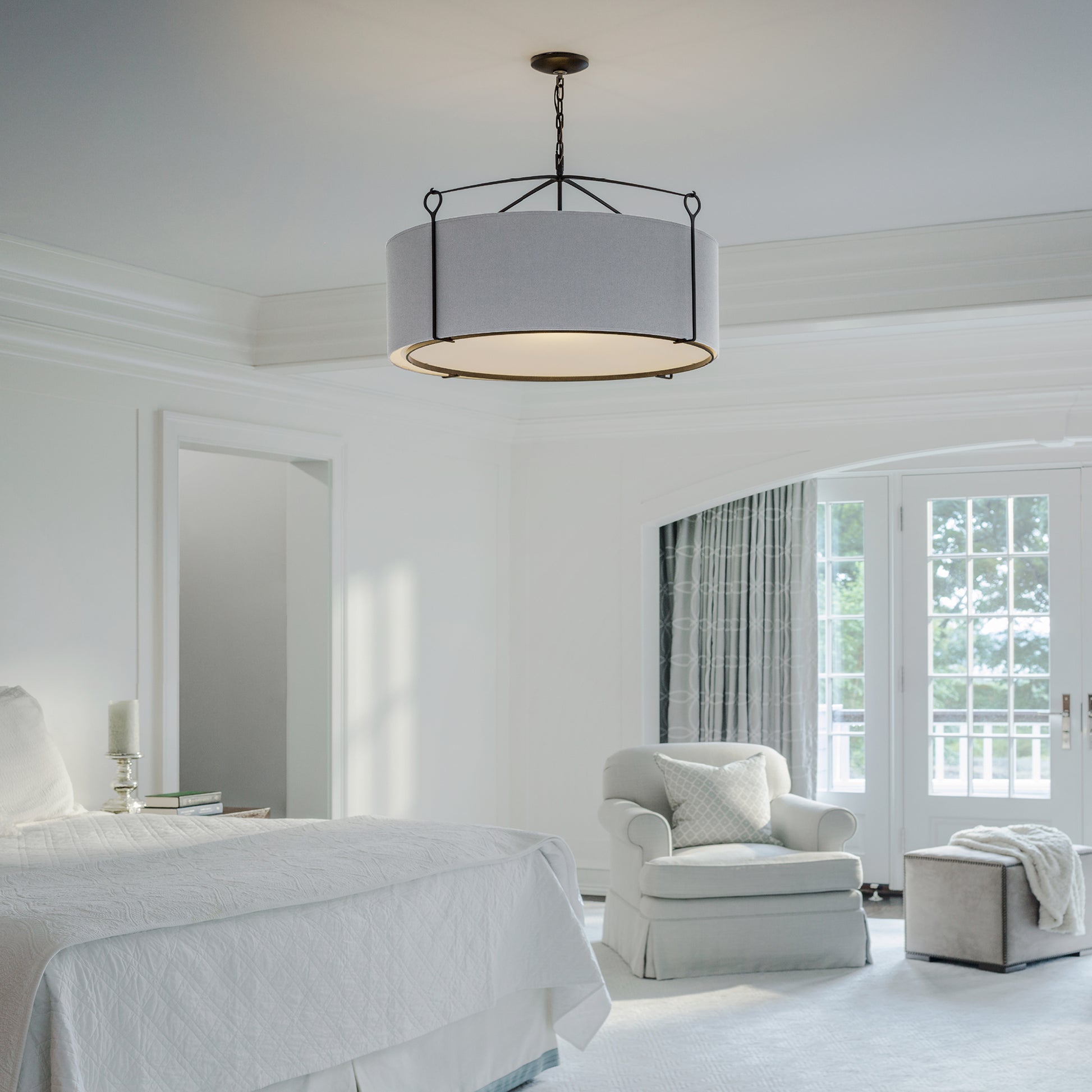 An elegant bedroom featuring white walls and linens, with a large window, an upholstered chair, a side table, and a Hubbardton Forge Bow Large Pendant ceiling lamp. The room is designed with a lavish yet