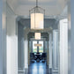A bright and airy hallway in a modern building featuring elegant Hubbardton Forge Tall Bow Pendant chandeliers with a soft glow hanging from the ceiling, leading to a room with visible window light and chairs.