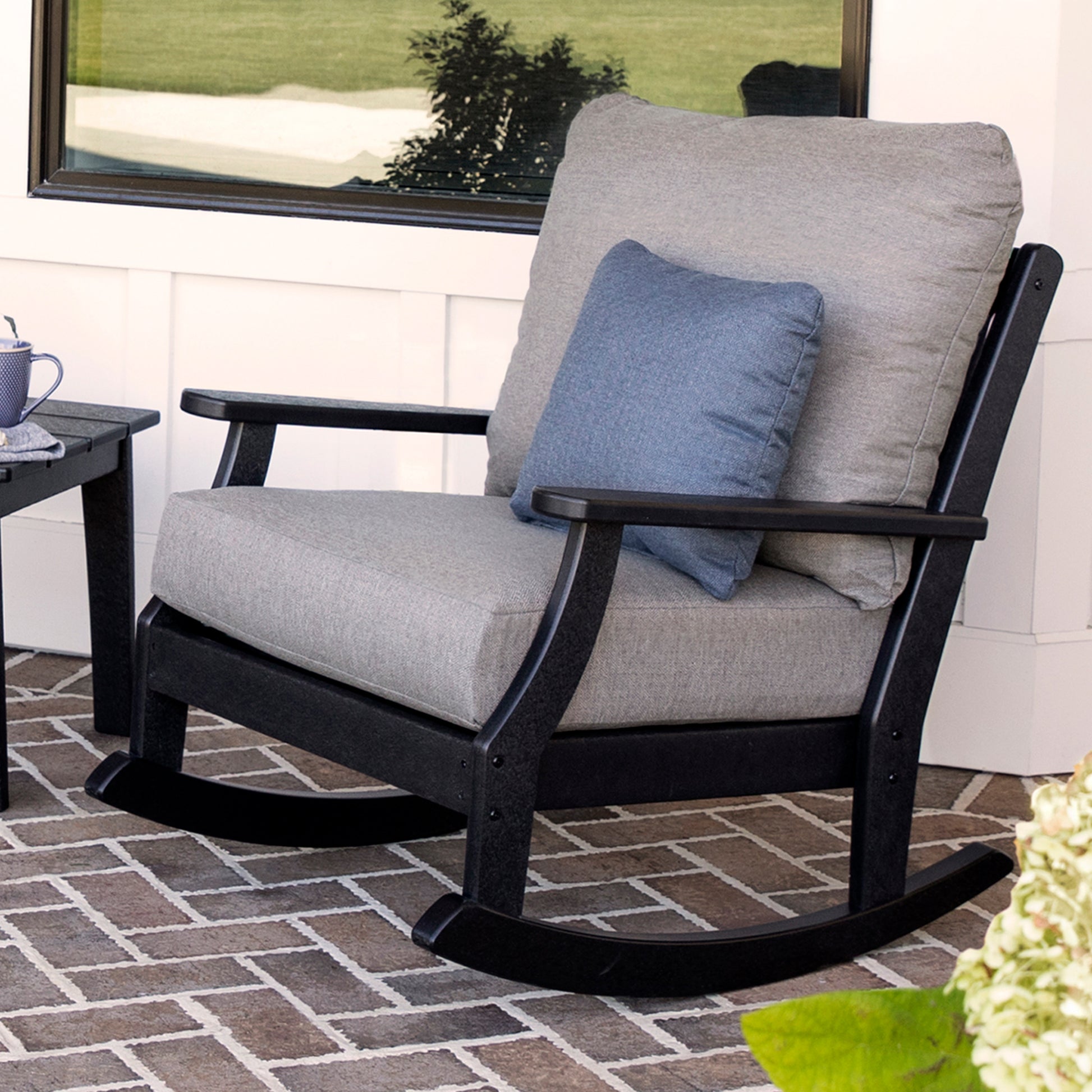 A gray-cushioned POLYWOOD Braxton Deep Seating Rocking Chair with a smaller blue pillow sits on a brick-patterned patio. A hint of a black coffee table and potted plant enhances the charm, while the window reflects lush greenery in the background, making it ideal for outdoor seating.