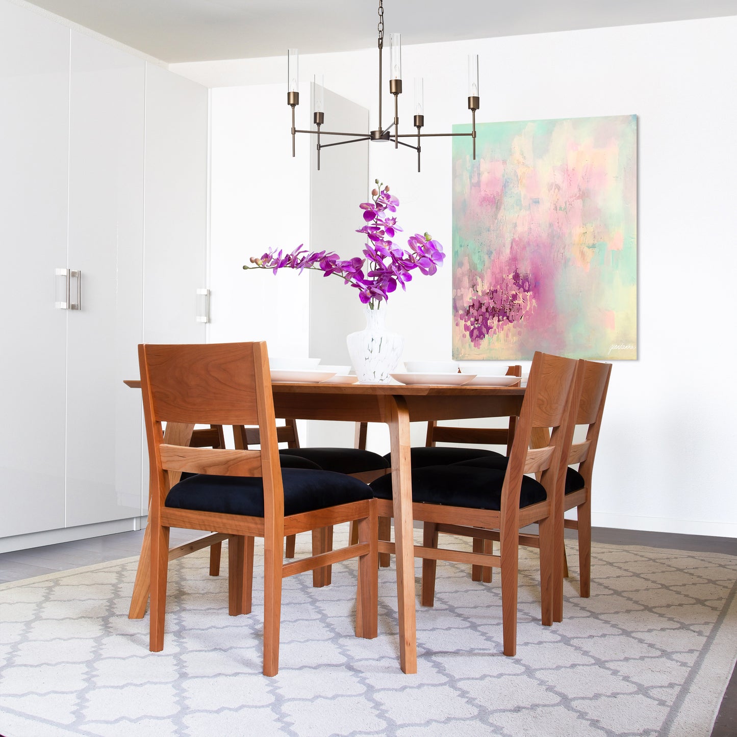 A modern dining room with a Vermont Woods Studios Burke Modern chair, a natural solid wood table set for four, dark cushioned chairs, a contemporary chandelier above, and an abstract painting on the wall.