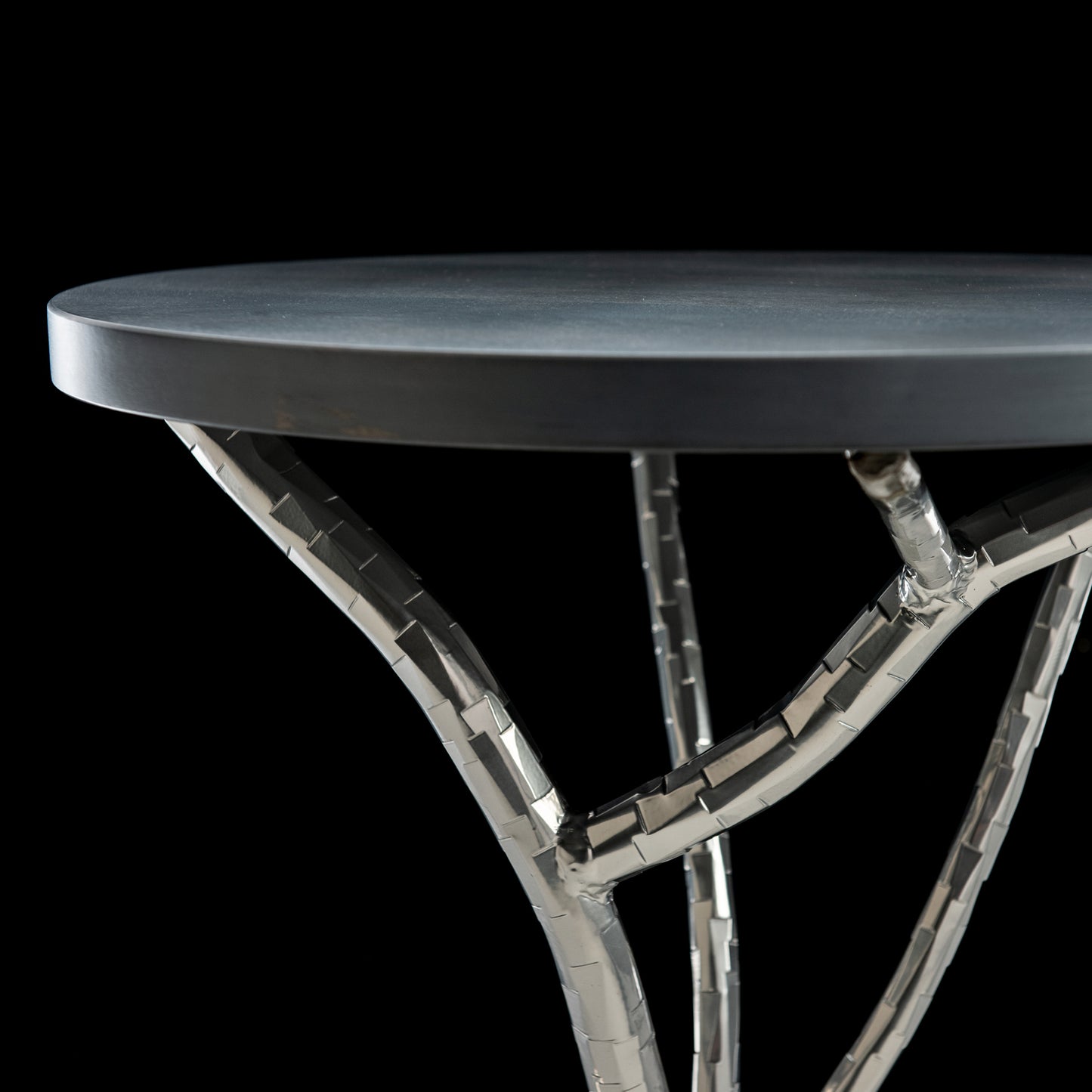 Close-up of the Brindille Wood Top Accent Table by Hubbardton Forge, featuring a round, dark tabletop and a metallic base. The intertwined metal branches create an artistic design reminiscent of Vermont maple trees. A solid black background accentuates the table's contemporary structure.