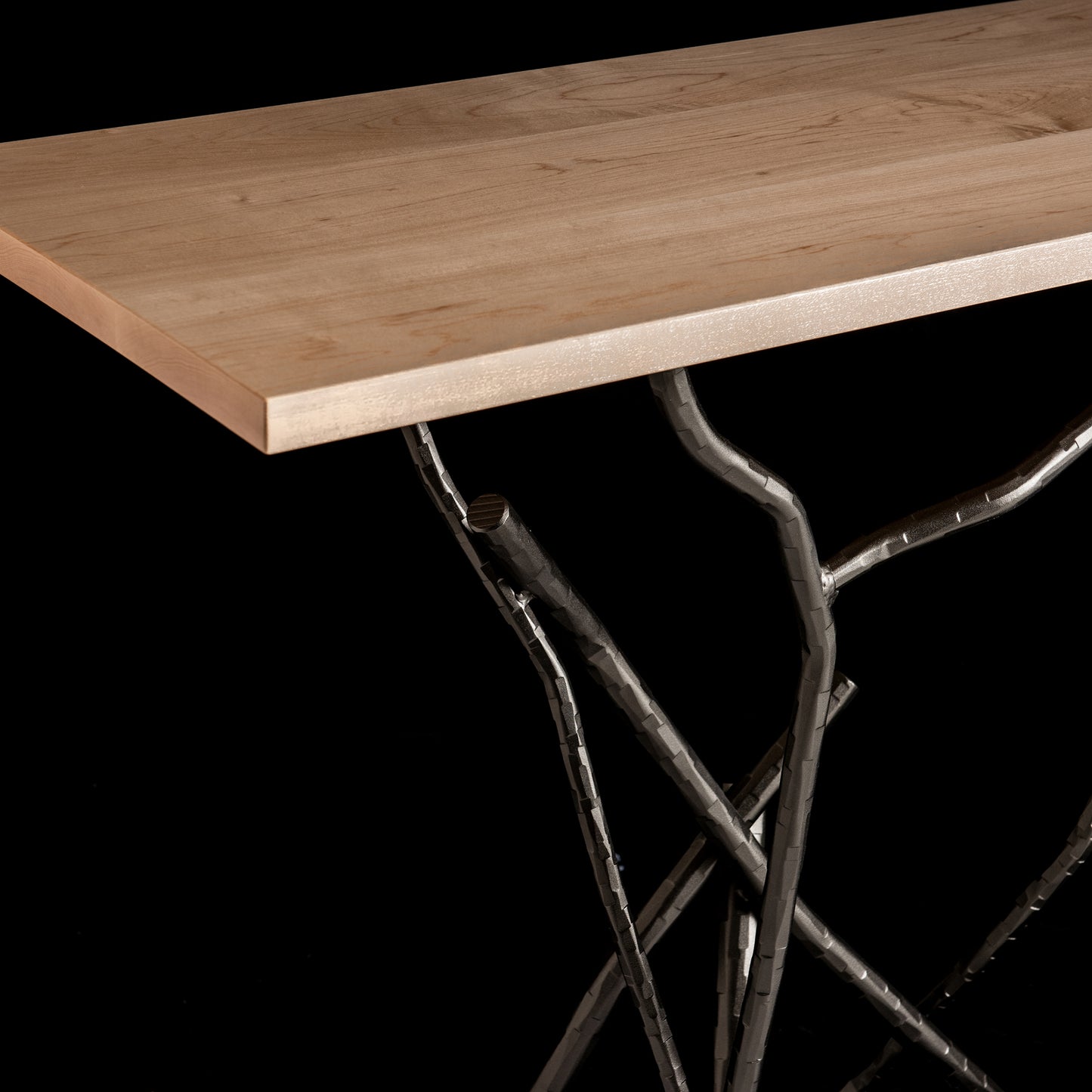 The Brindille Wood Top Console Table by Hubbardton Forge showcases a wooden tabletop with a sleek finish, resting on an artistically designed metal base featuring a twisted, branching motif. The solid black background highlights both the wood's natural texture and the table's intricate designer details.