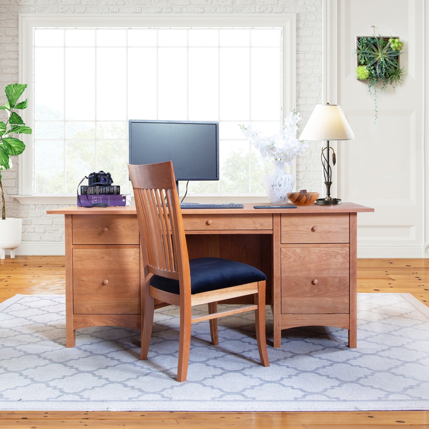 A handmade Vermont Furniture Designs Burlington Shaker Executive Desk and chair set in a bright room with a computer monitor, decorative plants, a camera, and various desk accessories.