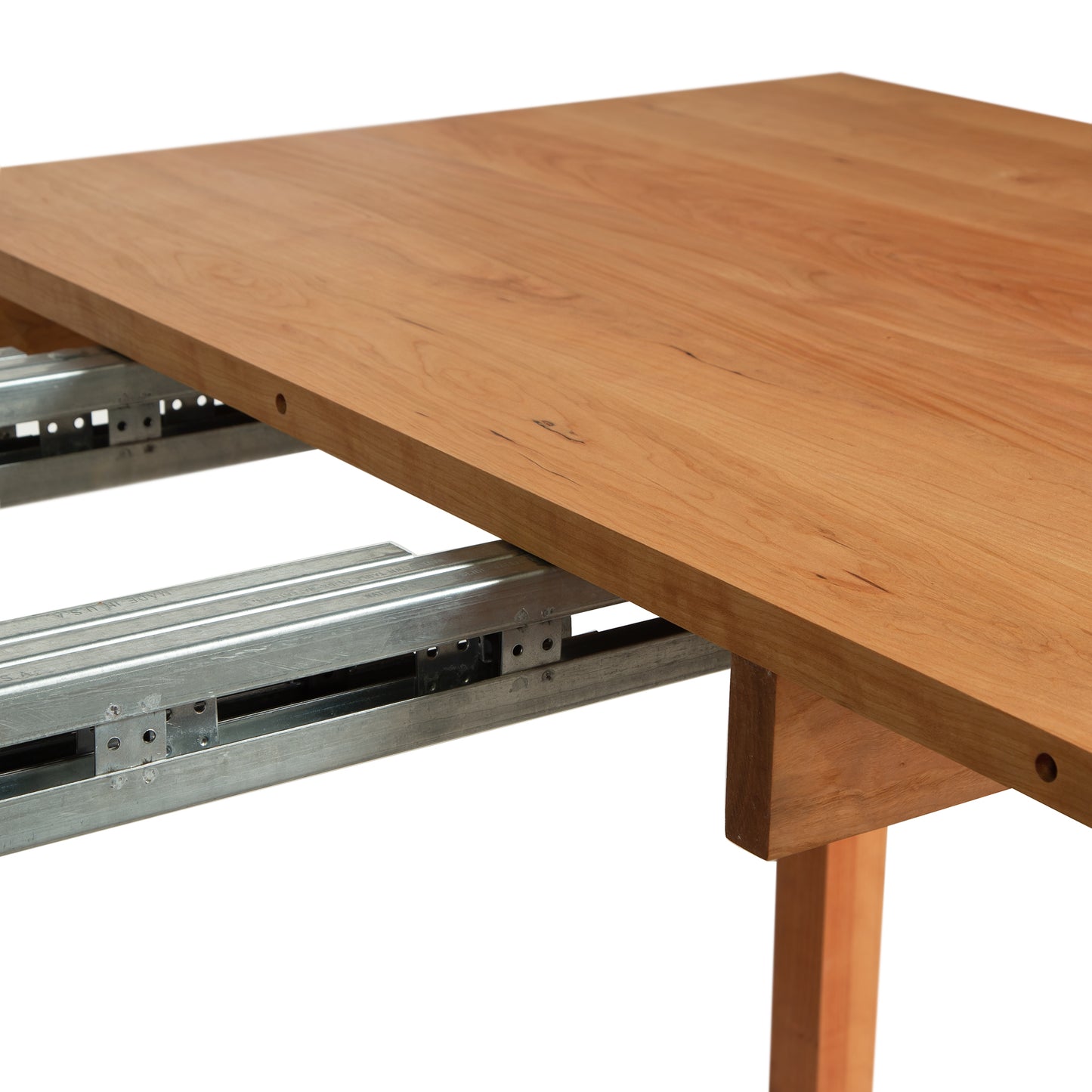 A close-up view of a Burlington Shaker Extension Dining Table - 40"x60" - Floor Model by Vermont Furniture Designs, shows the metal extension slides partially visible. Crafted by Vermont furniture makers, the table appears unfinished, and the extension mechanism is pulled out, showcasing the metal tracks beneath the wooden surface.