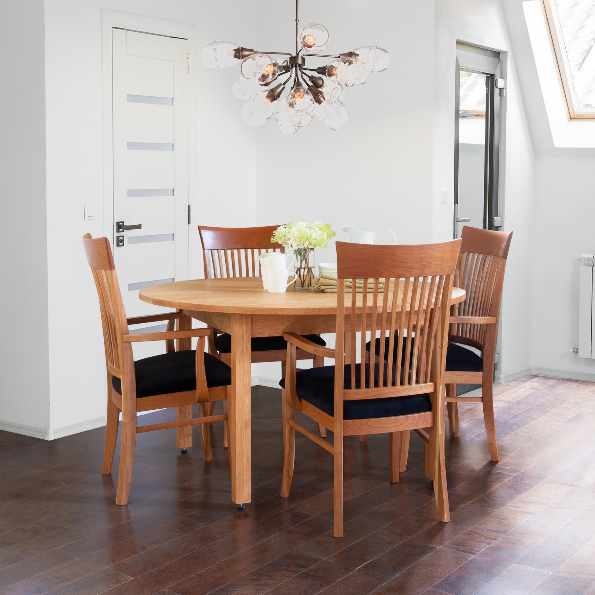 A modern dining room featuring a handcrafted Vermont Furniture Designs Burlington Shaker Round Solid Top Dining Table with six chairs, a decorative light fixture above, and flowers in a vase on the table, against a backdrop of white walls and a slo