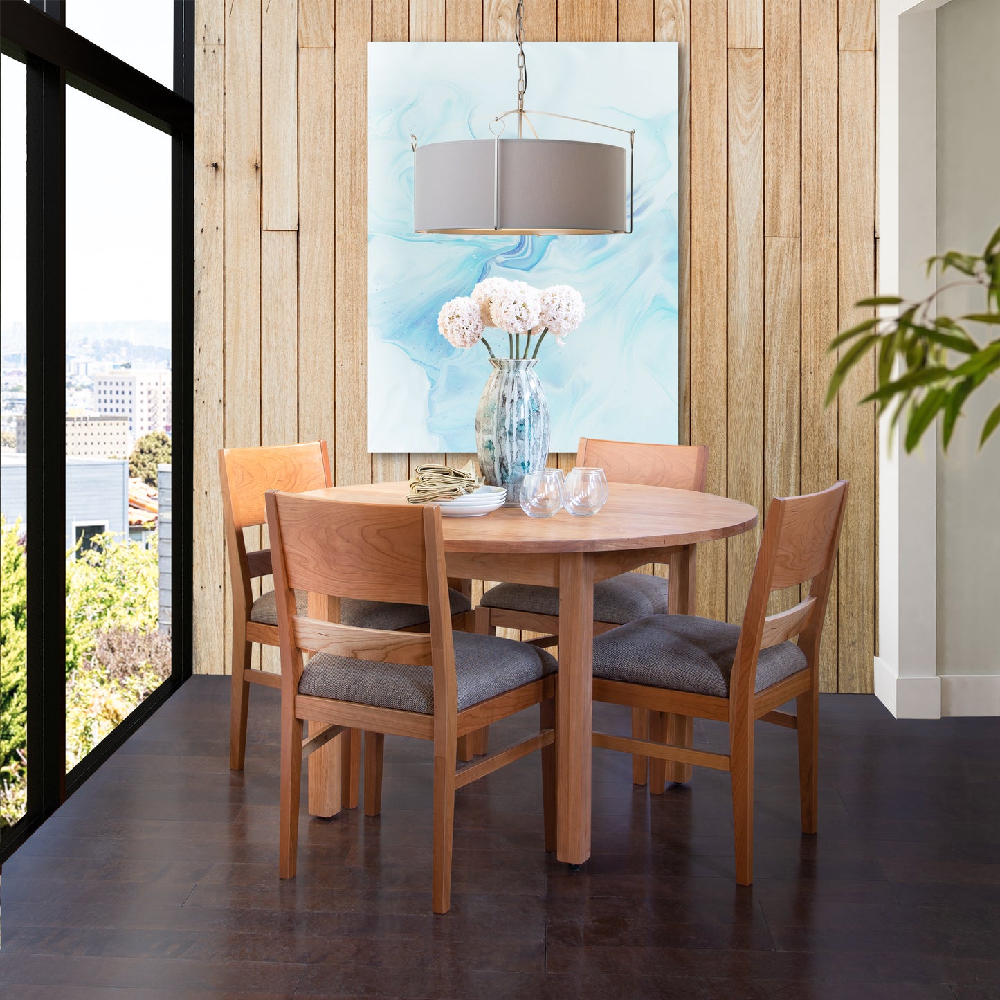 A modern dining area featuring a Vermont Furniture Designs Burlington Shaker Round Solid Top Dining Table with matching chairs, set against a backdrop of a cityscape view and a large blue art piece above the table.