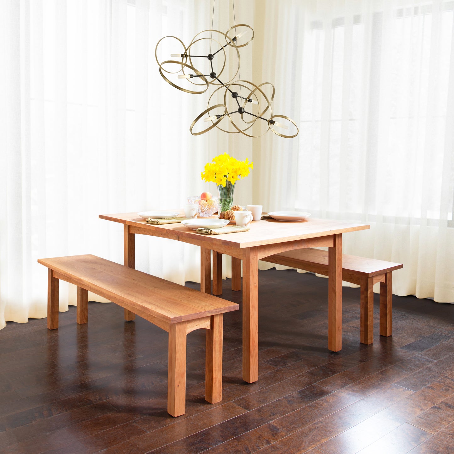 A wooden dining table set from the Vermont Furniture Designs Burlington Shaker Bench Collection, with two Vermont Furniture Designs Burlington Shaker Benches, place settings for two, and a bouquet of yellow flowers under a modern chandelier in a room with
