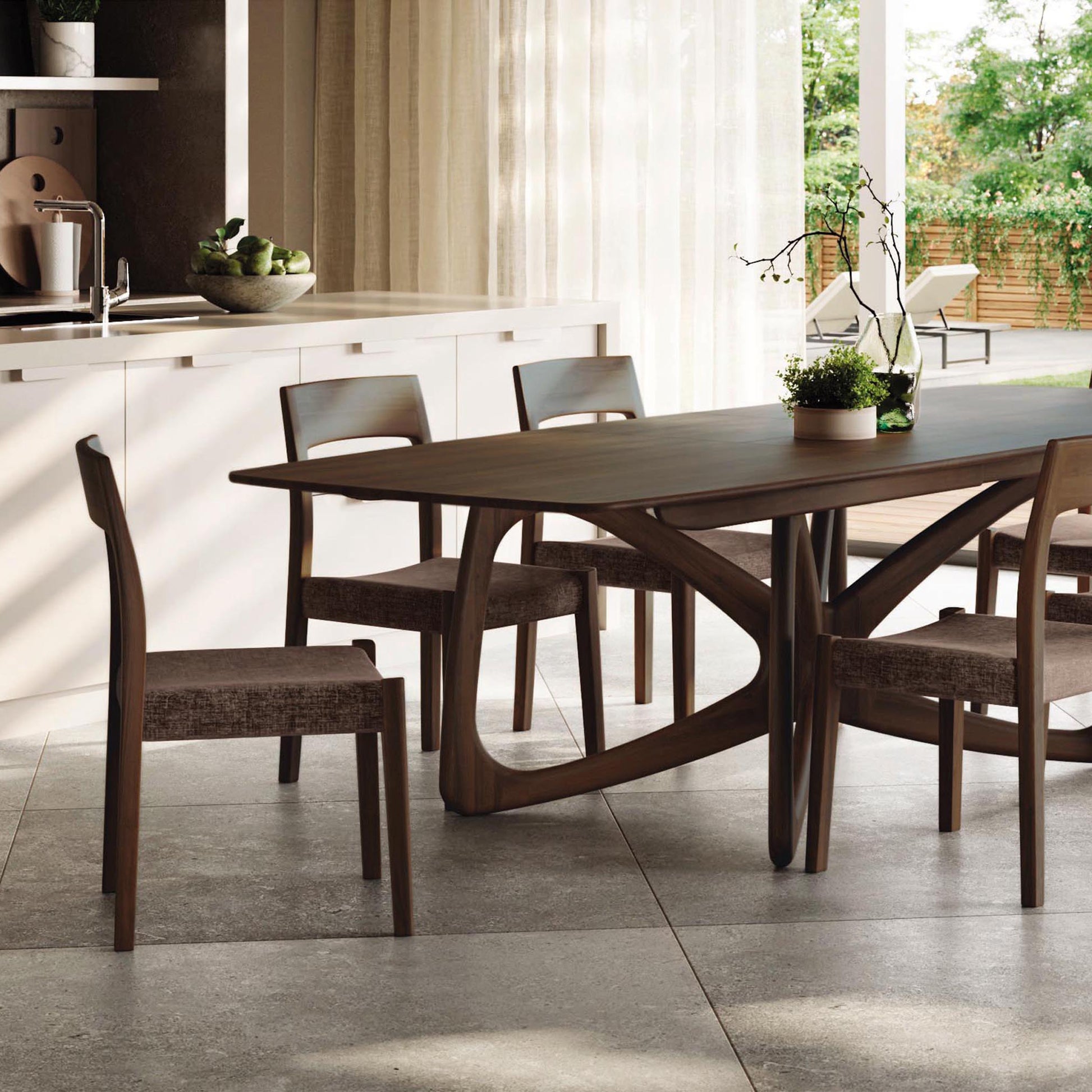 Alt text: A modern dining room with a sustainably sourced hardwood Butterfly Extension Dining Table by Copeland Furniture, accompanied by six cushioned chairs. The table's artistic base holds a small potted plant centerpiece. Large windows with sheer curtains allow natural light to flood the solid wood furniture setting.