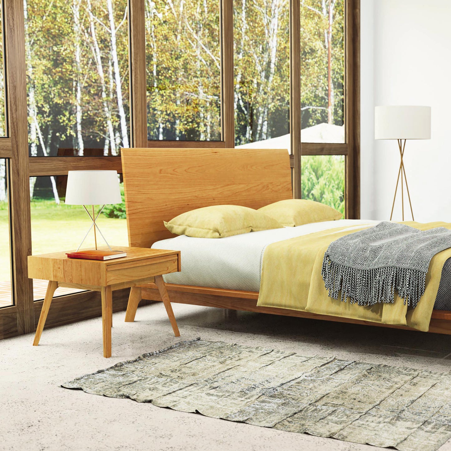 A bedroom features a wooden bed with yellow pillows and a gray blanket. A small solid wood Copeland Furniture Catalina 1-Drawer Nightstand with a lamp is next to the bed. The room has large floor-to-ceiling windows showing a view of trees. A standing lamp and a textured rug are also present, showcasing the beauty of sustainably harvested woods.