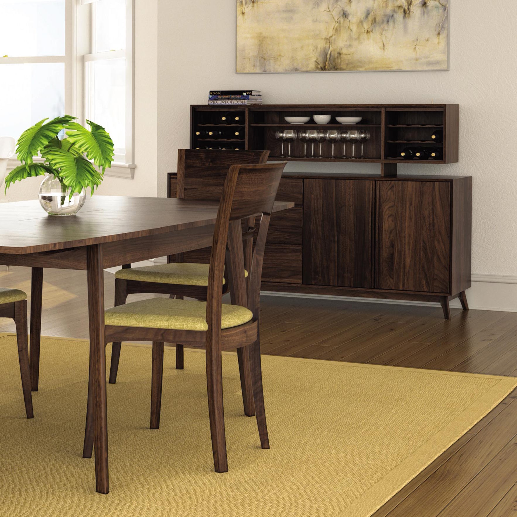 A dining room features a dark wood table with four chairs, each with light green cushions. The Copeland Furniture Catalina 3-Drawers, 2-Door Buffet, reminiscent of mid-century modern design, holds glasses in a rack and has a few books on top. A plant in a glass bowl sits on the table, and a painting hangs above the buffet.