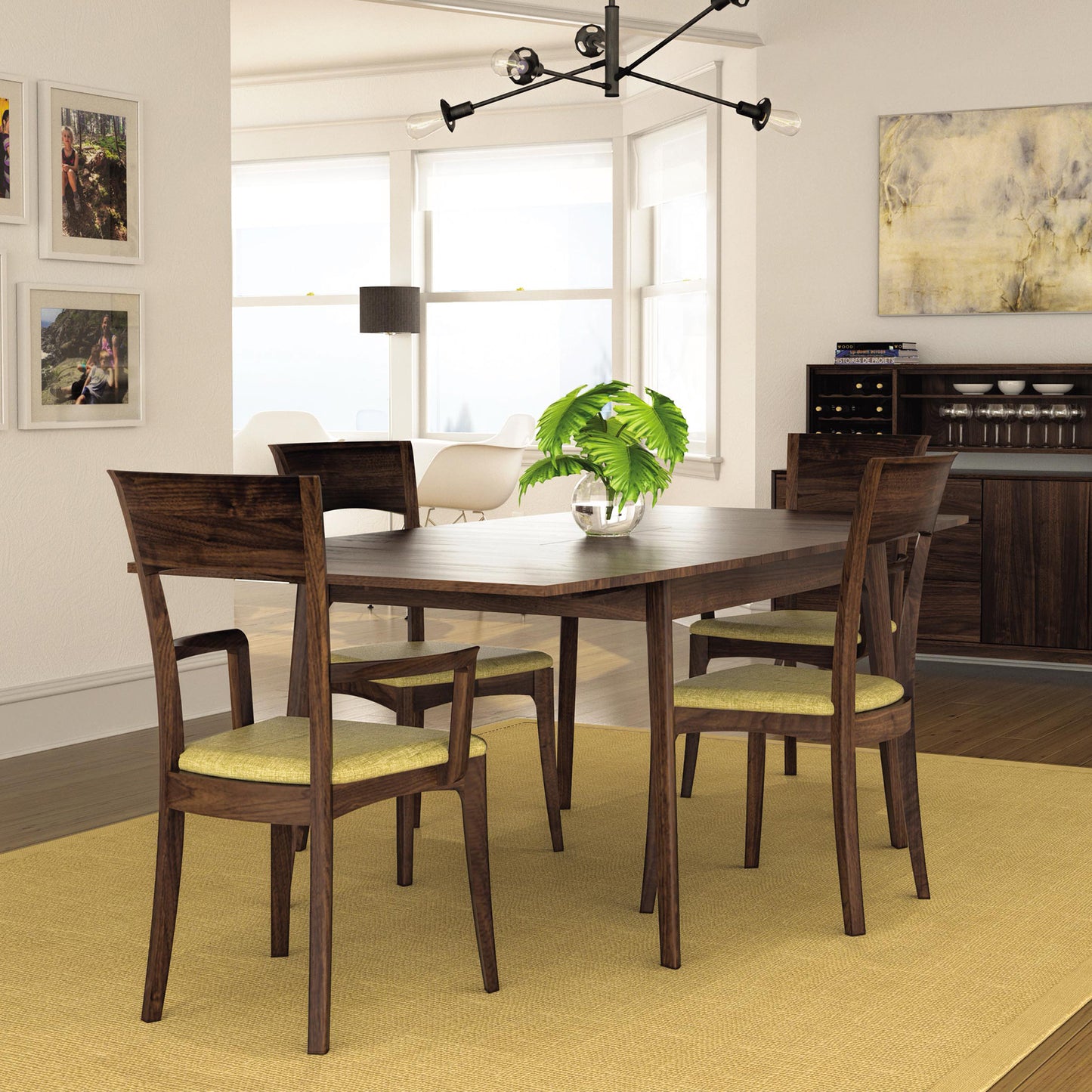 A dining room showcases a Copeland Furniture Catalina Extension Table made of solid wood, accompanied by six matching chairs with yellow cushions. The room is adorned with Mid-Century wall art, a modern light fixture, a plant centerpiece, and large windows. In the background, you can see a sideboard featuring a wine rack and additional furniture pieces.