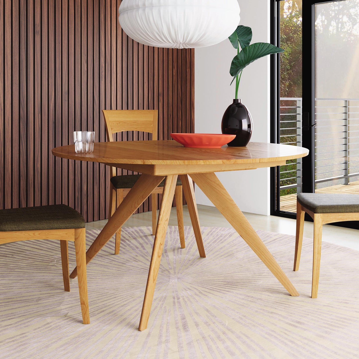 A modern dining area featuring the Copeland Furniture's Catalina Round Extension Table with three chairs. The round table is adorned with a red bowl and a dark vase with a green leafy plant. A white pendant light hangs above. The background shows a wooden slat wall and large window with a view outside.