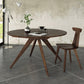 A custom handmade Catalina Round Extension Table by Copeland Furniture, featuring solid wood and angled legs, is positioned in the center of the room. A matching wooden chair sits next to it. The table holds a plant in a metal vase, a book, and a small decorative item. The room features a concrete floor and textured walls, adding to its modern charm.