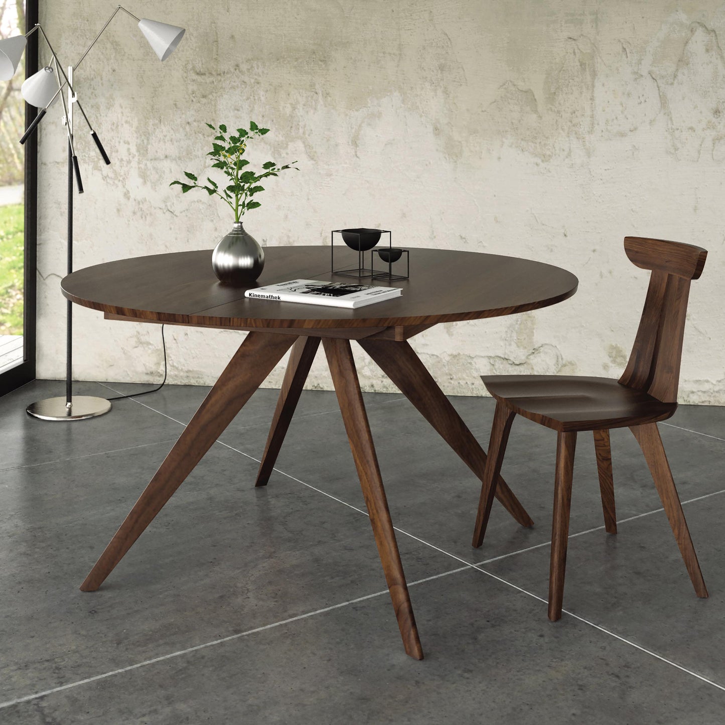 A custom handmade Catalina Round Extension Table by Copeland Furniture, featuring solid wood and angled legs, is positioned in the center of the room. A matching wooden chair sits next to it. The table holds a plant in a metal vase, a book, and a small decorative item. The room features a concrete floor and textured walls, adding to its modern charm.