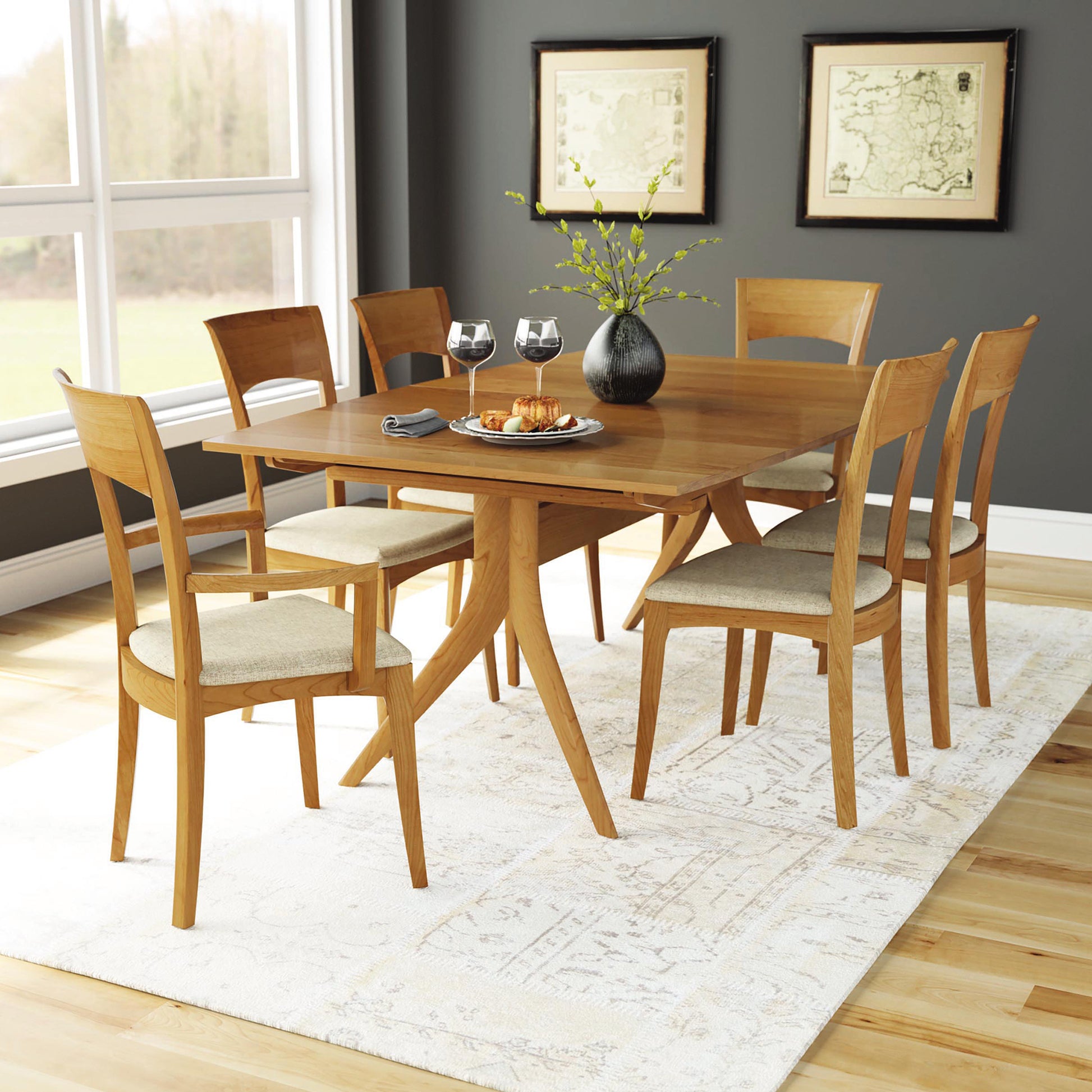 A modern dining room features a Copeland Furniture Catalina Trestle Extension Table made of solid American hardwood, accompanied by six matching chairs with cushioned seats. The table is set with appetizers, two wine glasses, and a vase with green branches. Large windows, a light-colored rug, and framed art complete the Mid-Century styling.