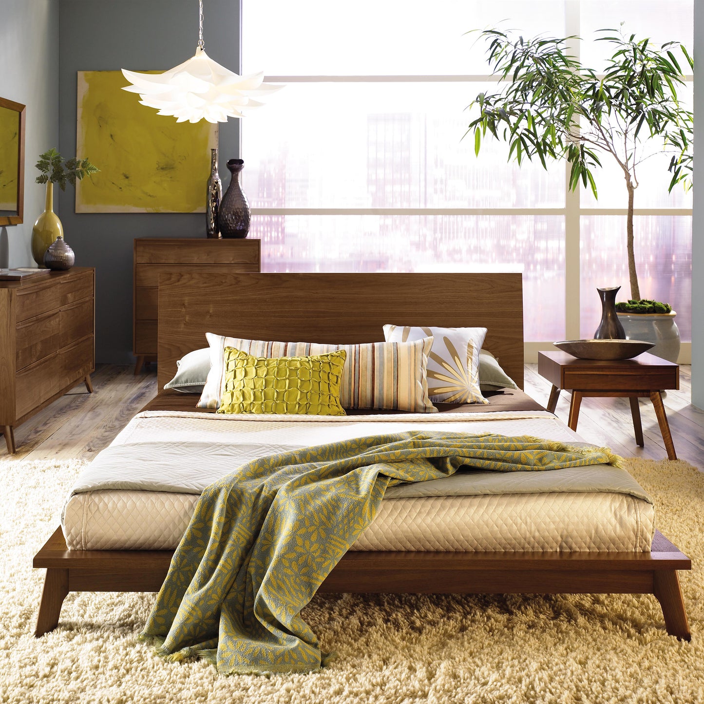 A modern bedroom showcases a Catalina Walnut Platform Bed by Copeland Furniture, dressed in white and green bedding. It is accompanied by a nightstand and dresser crafted from Vermont handmade furniture. A large potted plant sits to the right, while a contemporary light fixture hangs above. The room is flooded with natural light from the expansive windows in the background.