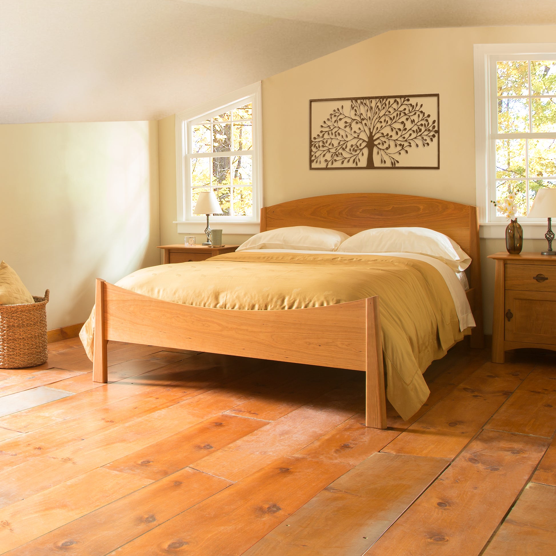 A bedroom with a wooden floor and the Maple Corner Woodworks' Cherry Moon Bed featuring beige bedding. Two windows above the headboard let in natural light, accentuating the tree artwork on the wall. Handmade solid wood nightstands sit on either side, enhancing the eco-friendly charm of this serene space.