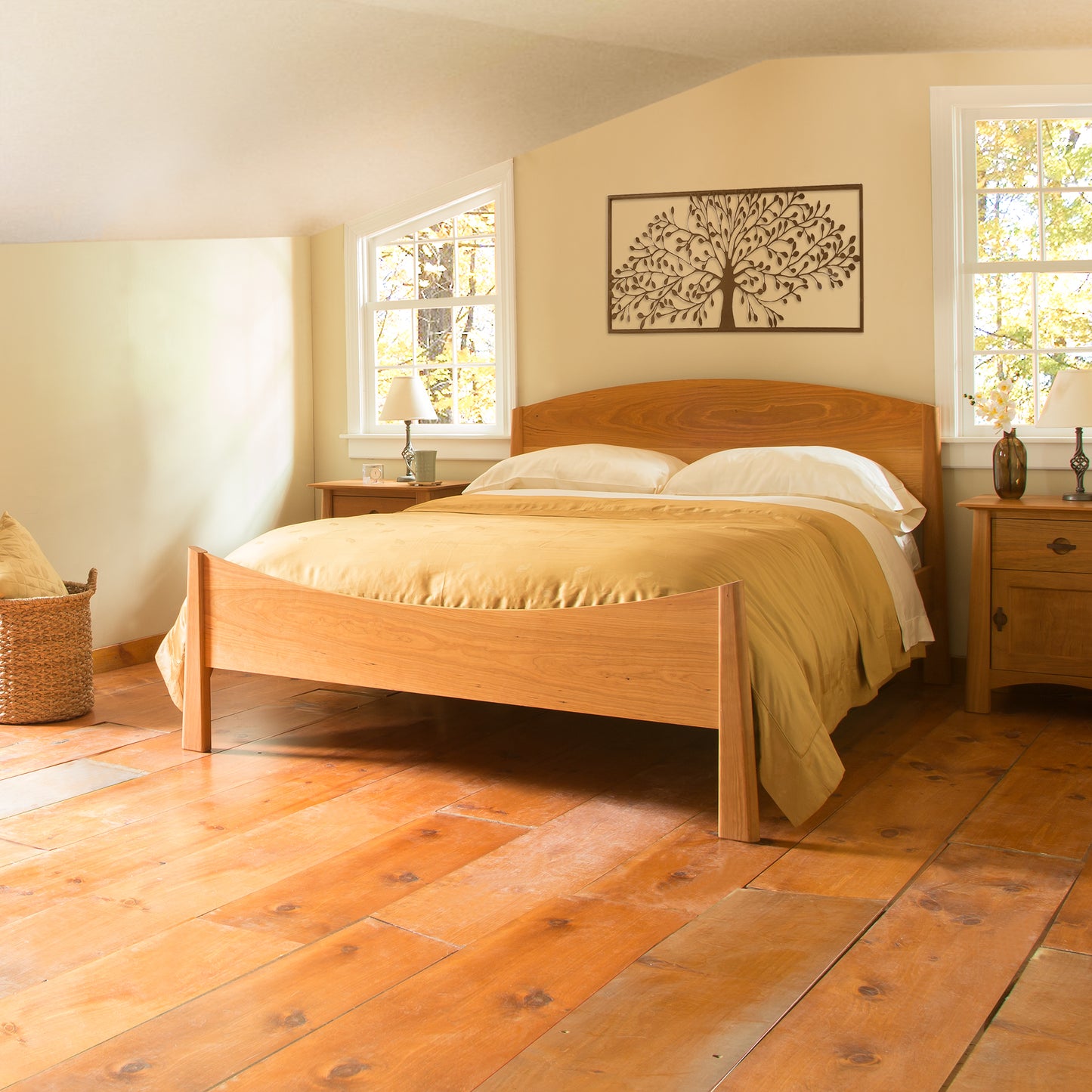 A warmly lit bedroom featuring a Maple Corner Woodworks Cherry Moon Bed with a yellow bedspread, polished wooden floor, and tree artwork above the bed. Two large windows let in sunlight, with wicker baskets in the corner.