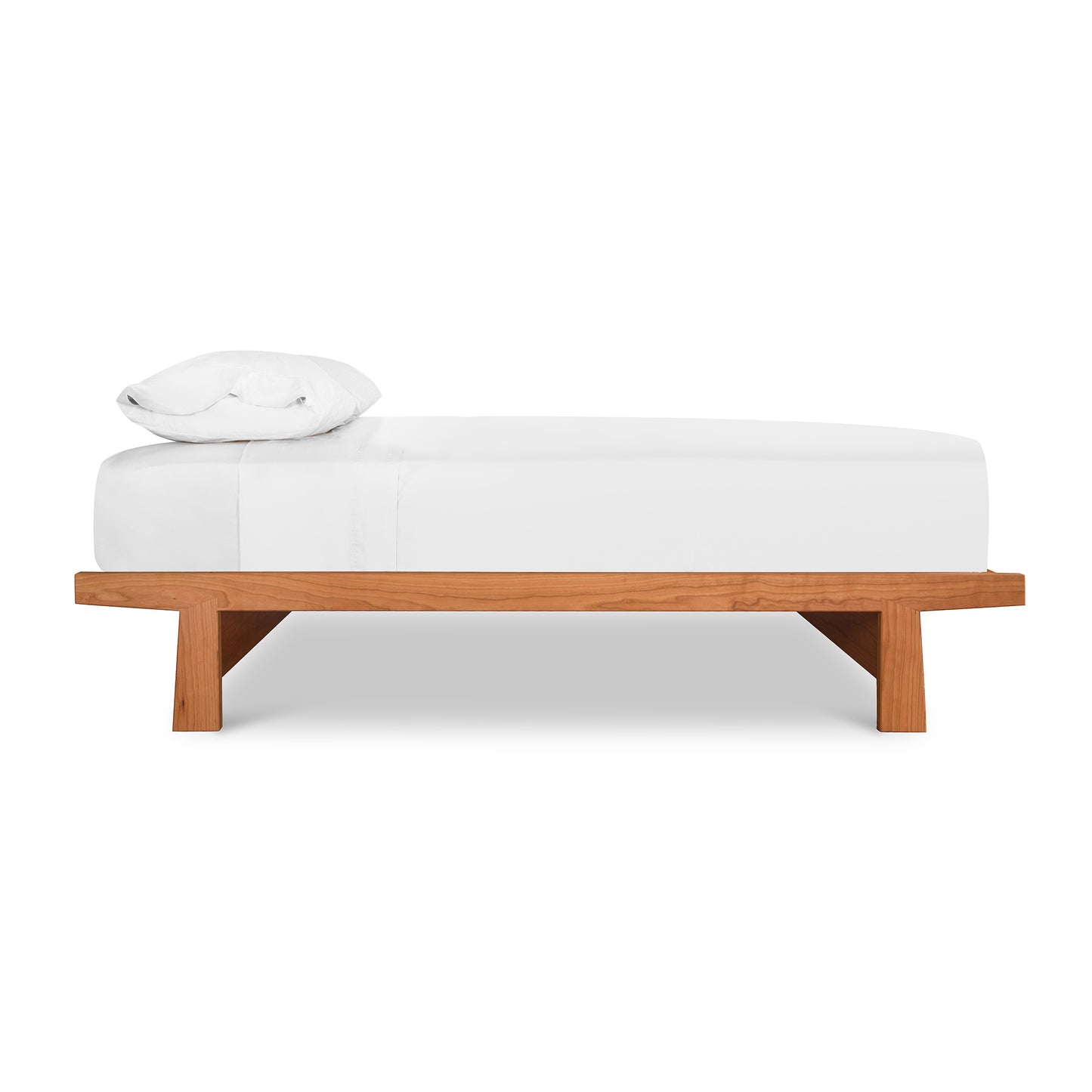 A simple modern Cherry Moon Dovetail Platform Bed with a wooden base crafted from solid, sustainably harvested woods and a white cushioned top, including a single white pillow at one end. The bed is set against a plain.