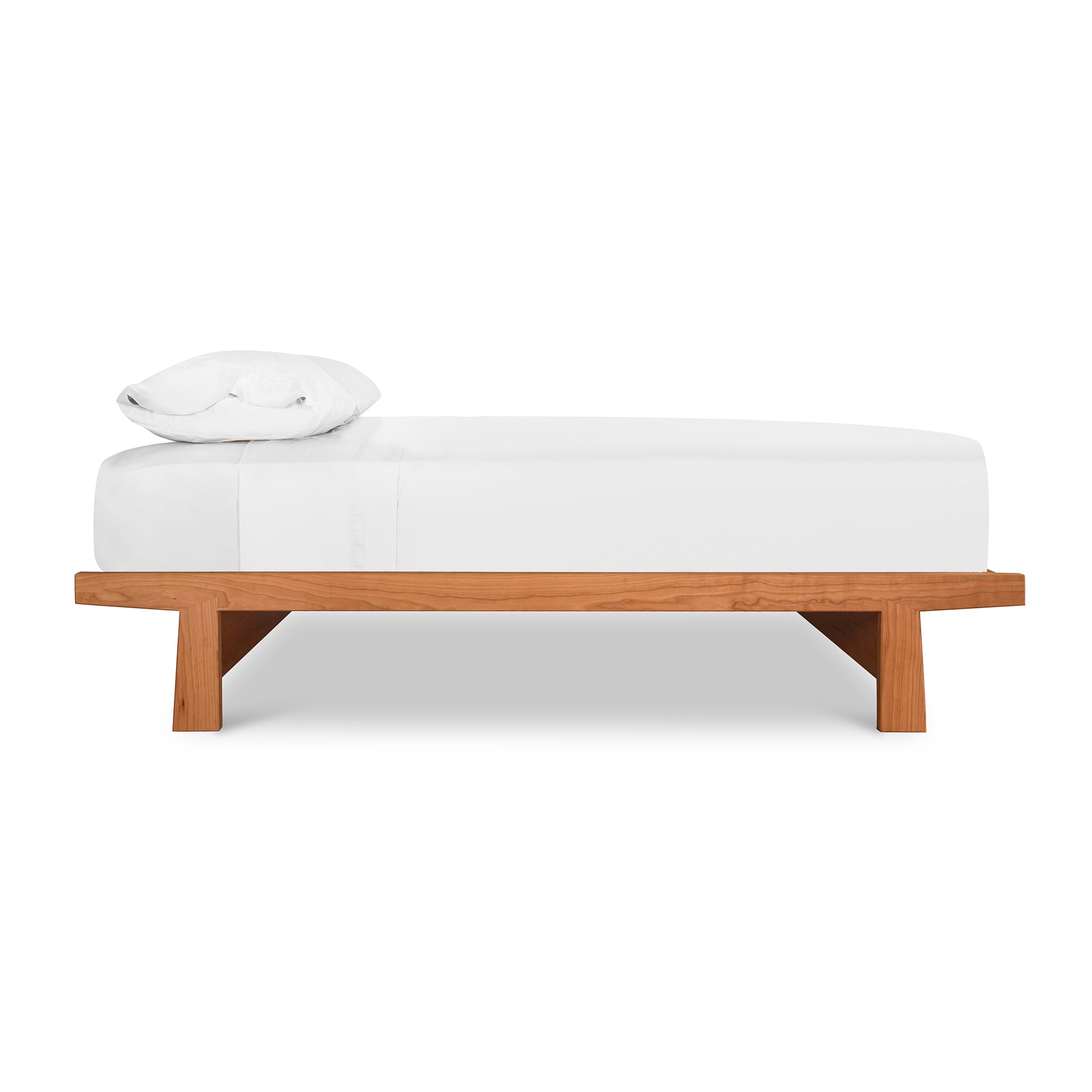 A simple modern Cherry Moon Dovetail Platform Bed with a wooden base crafted from solid, sustainably harvested woods and a white cushioned top, including a single white pillow at one end. The bed is set against a plain.