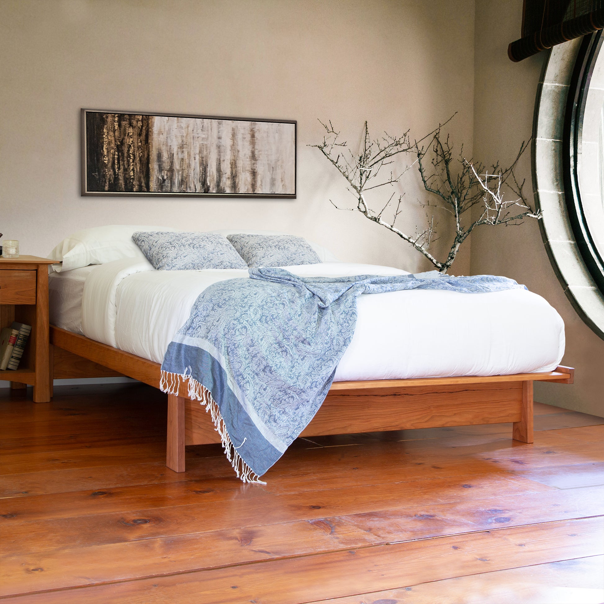 Cherry Moon Dovetail Platform Bed in a stylish bedroom featuring white bedding, blue patterned pillows, nightstand with books, and abstract wall art. Sustainable cherry wood furniture on wooden flooring. Perfect example of American made maple and cherry wood furniture by Maple Corner Woodworks.