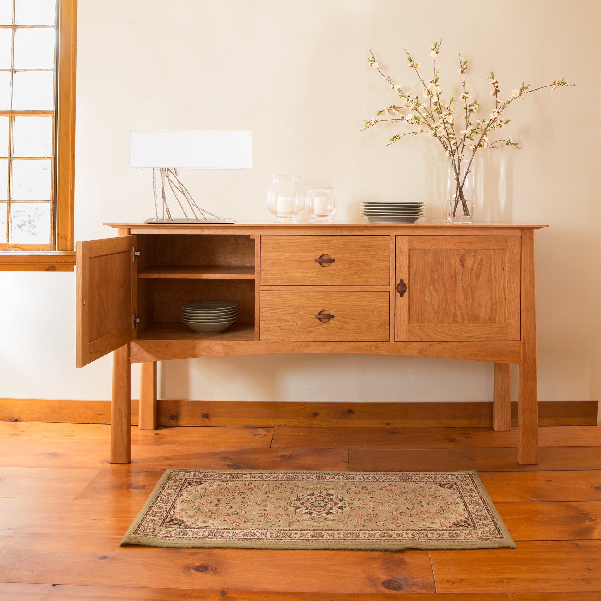 A Cherry Moon Huntboard from Maple Corner Woodworks stands elegantly on a polished wooden floor. It features two drawers and an open cabinet with a few plates, embodying an Asian style. On top, there's a small lamp, jars, and a vase with branches. A decorative rug lies in front, illuminated by the luxury kitchen window's glow.