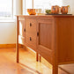 A Maple Corner Woodworks Cherry Moon Huntboard with two drawers and two cabinets, adorned with glass pitchers and a bowl of fruit, set against a sunny window in a room with natural hardwood floors.