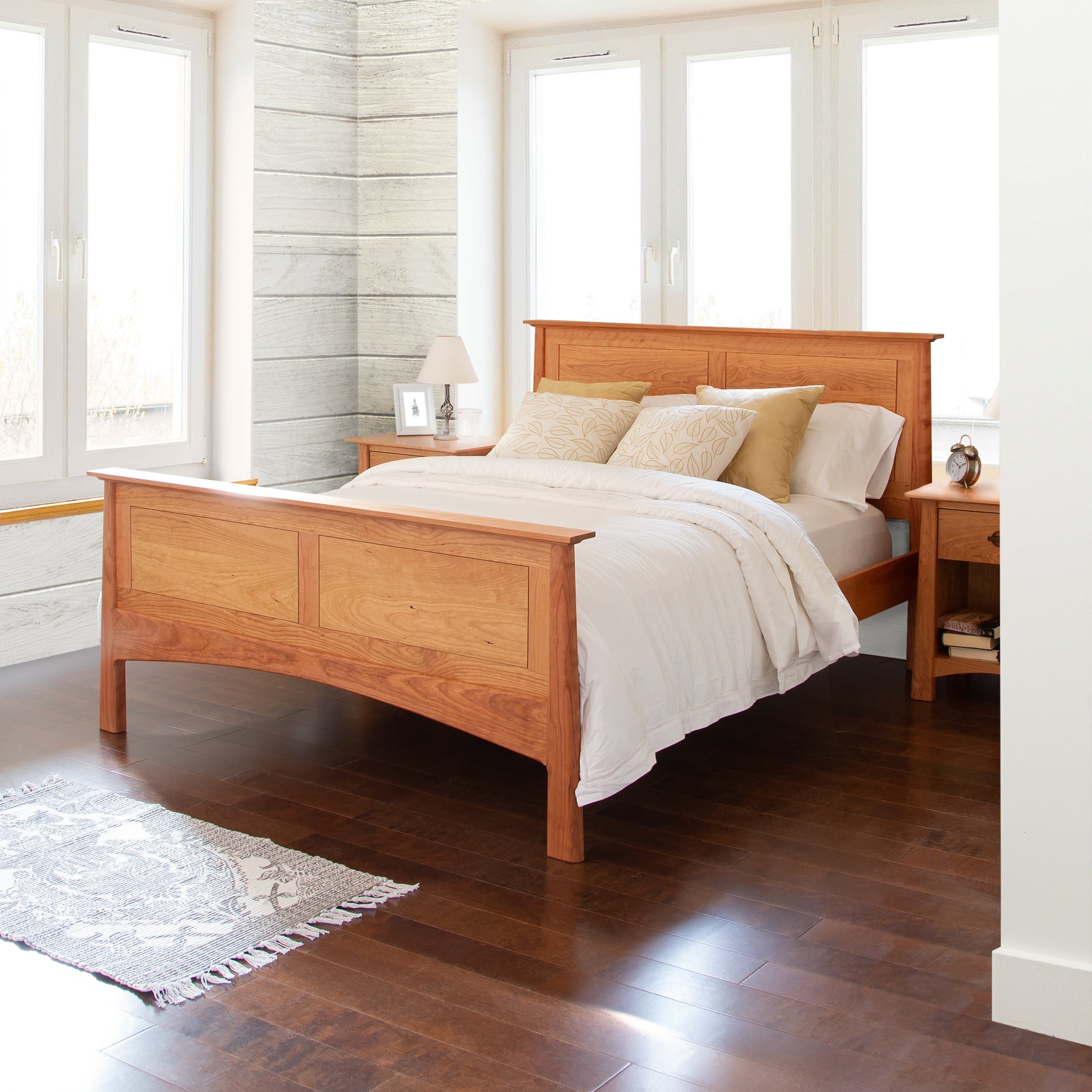 A Maple Corner Woodworks Cherry Moon Panel Bed with white bedding in a bright bedroom, featuring large windows, a gray rug, and a minimalist nightstand. The walls are white and a unique cylindrical gray stone column is visible.
