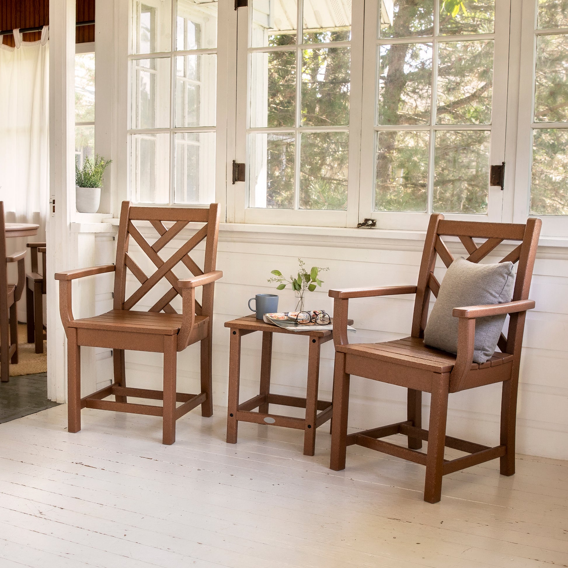 Two POLYWOOD Chippendale Outdoor Dining Arm Chairs with X-back designs are arranged on a white porch floor, placed on either side of a small table with a vase holding greenery. A gray cushion adorns one of the chairs. Large windows in the background provide views of flourishing greenery outside, highlighting the durable outdoor furniture.