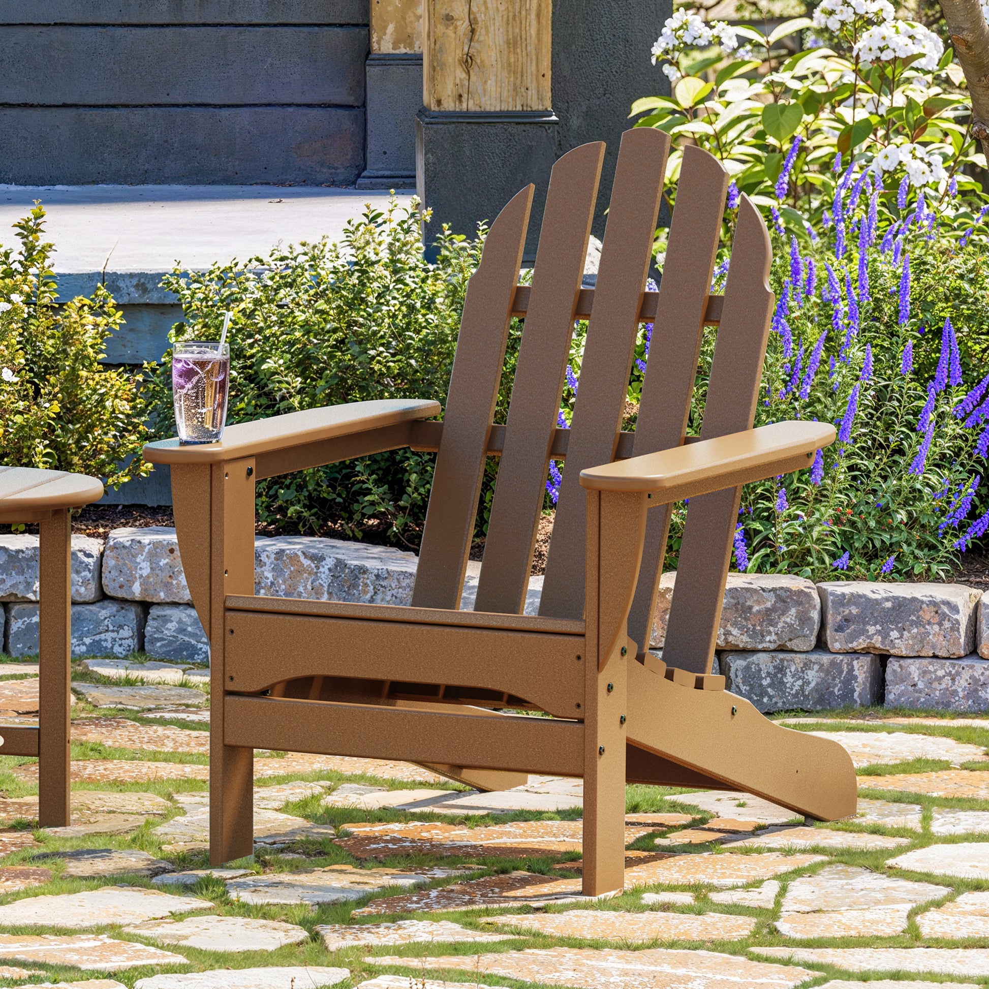 A POLYWOOD Classic Adirondack Chair, made from durable POLYWOOD lumber, sits on a stone patio. A glass with a beverage rests on its right armrest, highlighting its functionality. In the background, vibrant flowers and plants embellish a garden bed in front of a house.