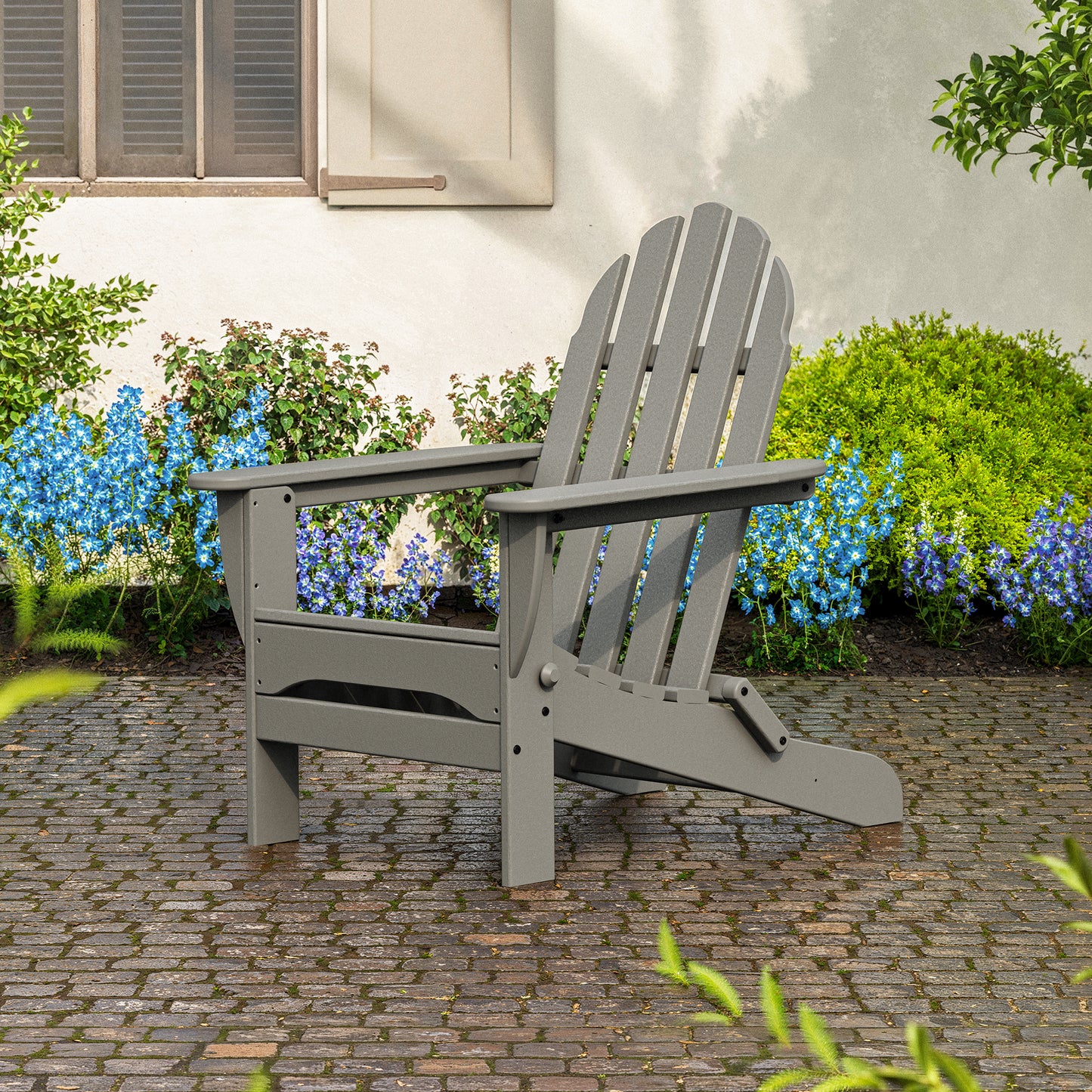 A POLYWOOD Classic Folding Adirondack Chair in gray, made from recycled plastic, is placed on a cobblestone patio in a garden setting. The backdrop showcases green bushes and clusters of small blue flowers against a white building wall with a shuttered window, creating an inviting piece of outdoor furniture.