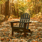 A POLYWOOD Classic Folding Adirondack Chair rests on a path covered with fallen autumn leaves. Trees with vibrant foliage line the path, creating a serene woodland scene under warm sunlight.