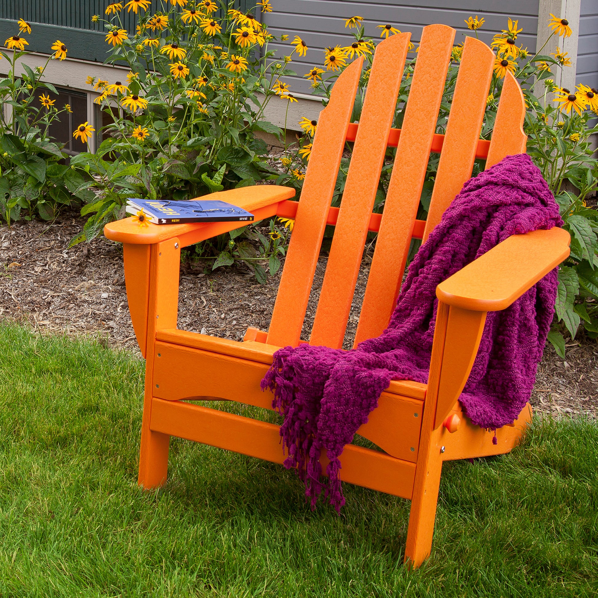 A POLYWOOD Classic Folding Adirondack Chair in orange, crafted from recycled plastic, sits on a lawn. A purple blanket is draped over it, and a closed book rests on the left armrest. Yellow flowers and the exterior wall of the house create a charming backdrop.