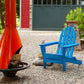 A brightly colored outdoor scene showcases a vivid blue POLYWOOD Classic Folding Adirondack Chair, made from recycled plastic, situated on a gravel patio. Next to it, a hat and cushion rest on the ground. An orange chiminea holds firewood, while a can is placed on the chair arm. A red and green building stands in the background.