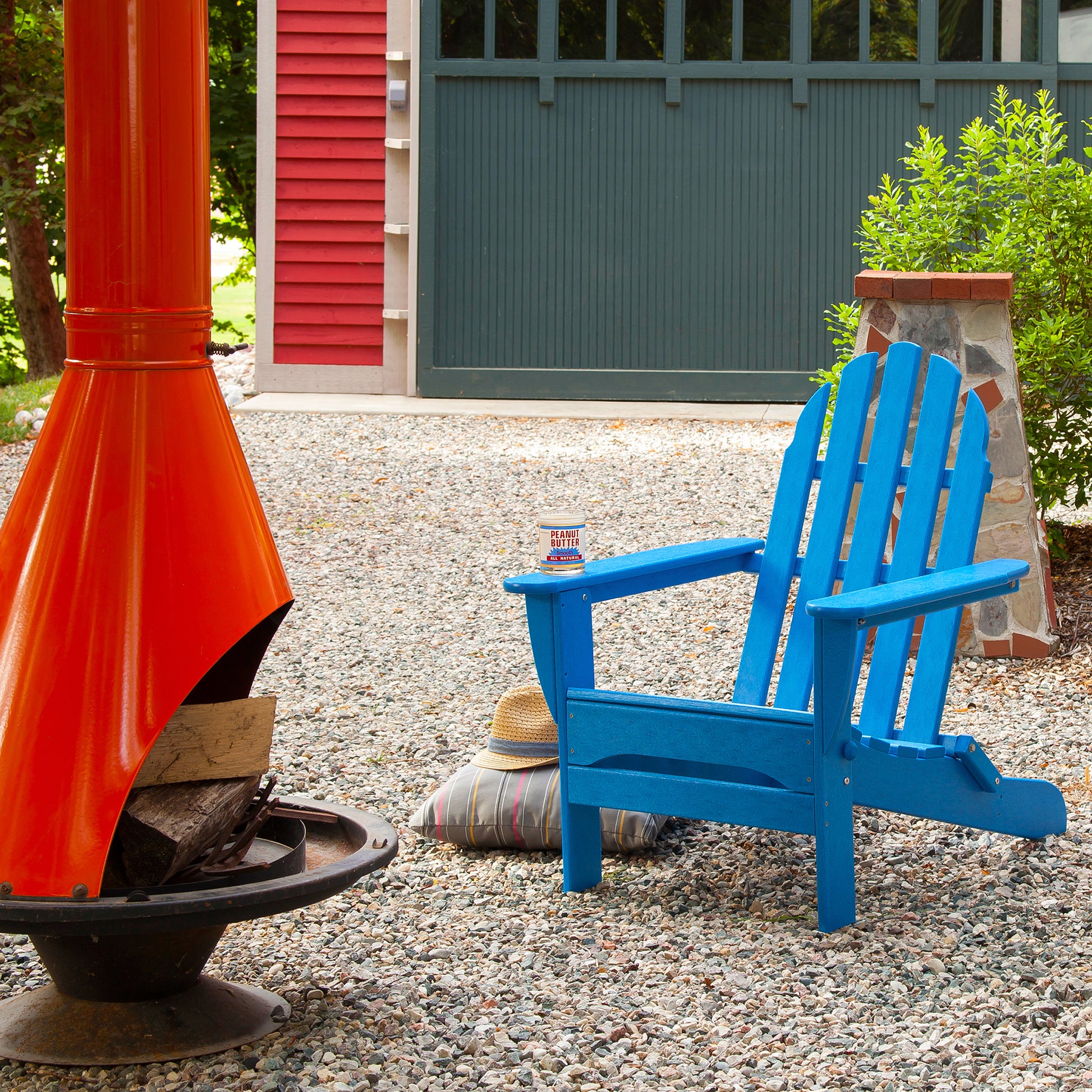 A brightly colored outdoor scene showcases a vivid blue POLYWOOD Classic Folding Adirondack Chair, made from recycled plastic, situated on a gravel patio. Next to it, a hat and cushion rest on the ground. An orange chiminea holds firewood, while a can is placed on the chair arm. A red and green building stands in the background.