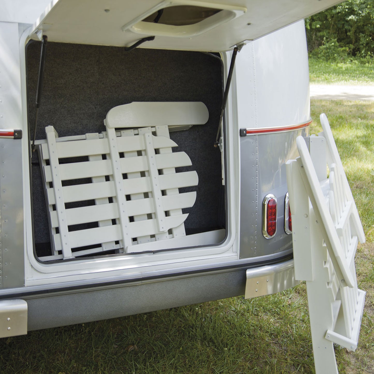 The image shows the rear storage compartment of a vehicle with a door open, revealing folded white wooden furniture, including a POLYWOOD Classic Folding Adirondack Chair. A length of white metal ladder leans against the opened door, set against a green grass background.