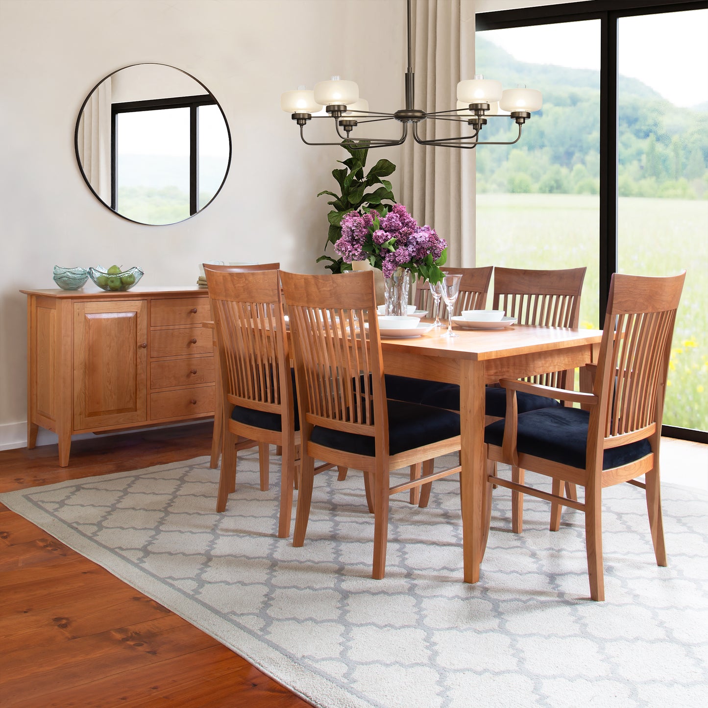 A well-lit dining room featuring a wooden table with six Vermont Woods Studios Contemporary Shaker chairs, a sideboard, and a circular mirror. Large windows show a scenic outdoor view. Decor includes a chandelier and