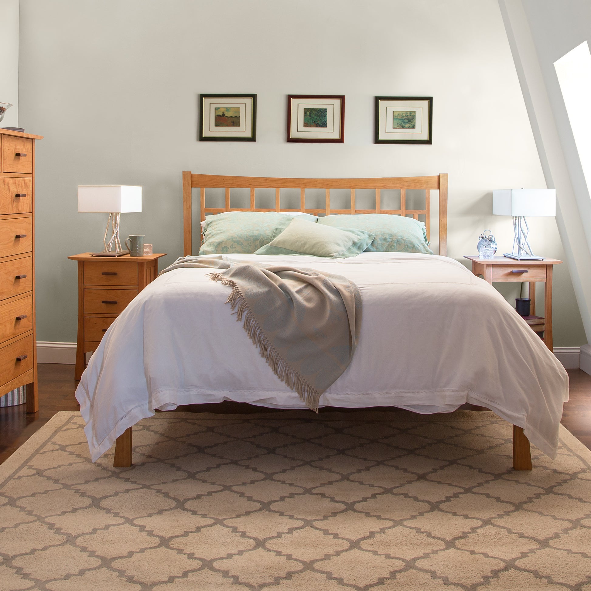 A neatly made solid wood bed from the Vermont Furniture Designs Contemporary Craftsman Low Footboard Bed collection with white bedding and a beige throw blanket in a room with angled walls, two bedside tables with lamps, and framed