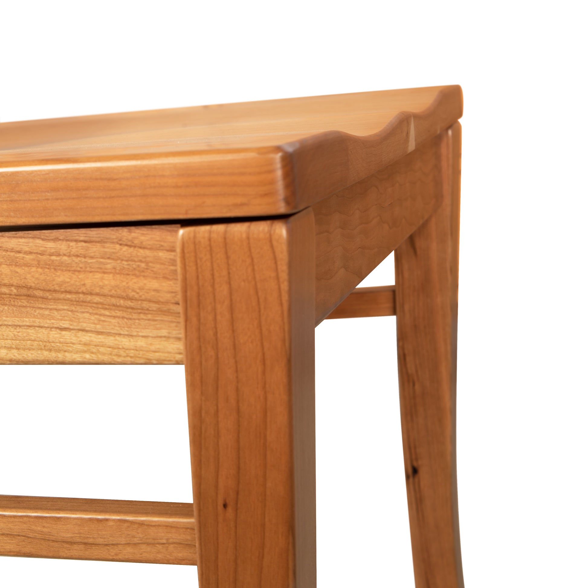 A close-up photo of a wooden table. The table has a smooth, polished finish with clean lines and sturdy legs. The corner of the table, made from natural cherry wood, is prominently shown against a plain white background. In the scene, the elegant Contemporary Shaker Side Chair with Wood Seat - Display Model by Vermont Woods Studios is subtly featured next to the table.