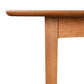 A close-up shot captures the corner of the Custom Heartwood Shaker Extension Dining Table, crafted from solid maple wood by Vermont Furniture Designs. The table features an eco-friendly oil finish that highlights the visible wood grain. Its tabletop, with slightly rounded edges, is supported by legs and a crossbeam intersecting at a right angle. The wood has a light, natural color.