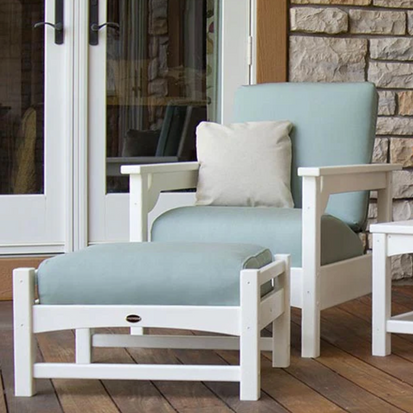 An outdoor seating area featuring a white wooden lounge chair with light blue cushions and a matching ottoman. The chair has a grey square pillow. The background shows a stone wall and a glass door.