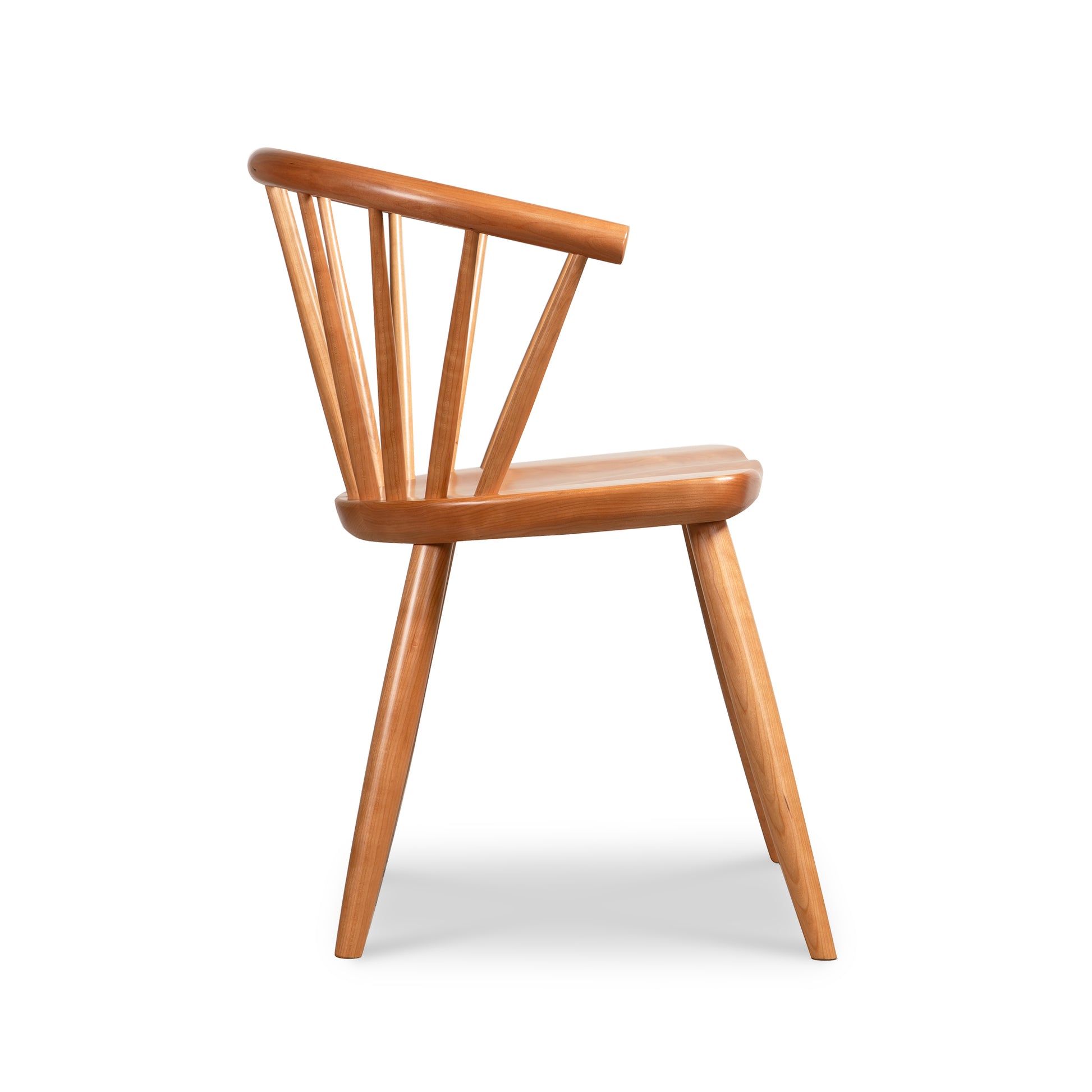 A side view showcases the Dover Curveback Windsor Chair by Vermont Woods Studios, highlighting its wooden structure with a light brown finish. The chair features a rounded backrest with vertical slats and slightly angled legs, while its minimalist seat and overall design provide a contemporary twist on traditional Windsor-style elegance.