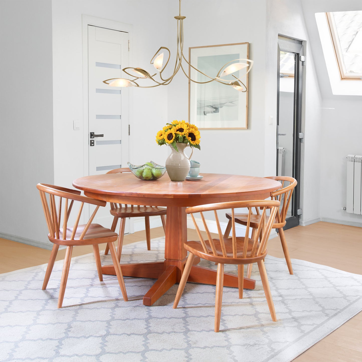 A dining room with a wooden round table, four matching Dover Curveback Windsor Chairs from Vermont Woods Studios featuring contoured wood seats, and a vase of sunflowers on the table. A bowl of green apples is also on the table. There's a modern chandelier above, a door in the background, and a bright, airy ambiance.