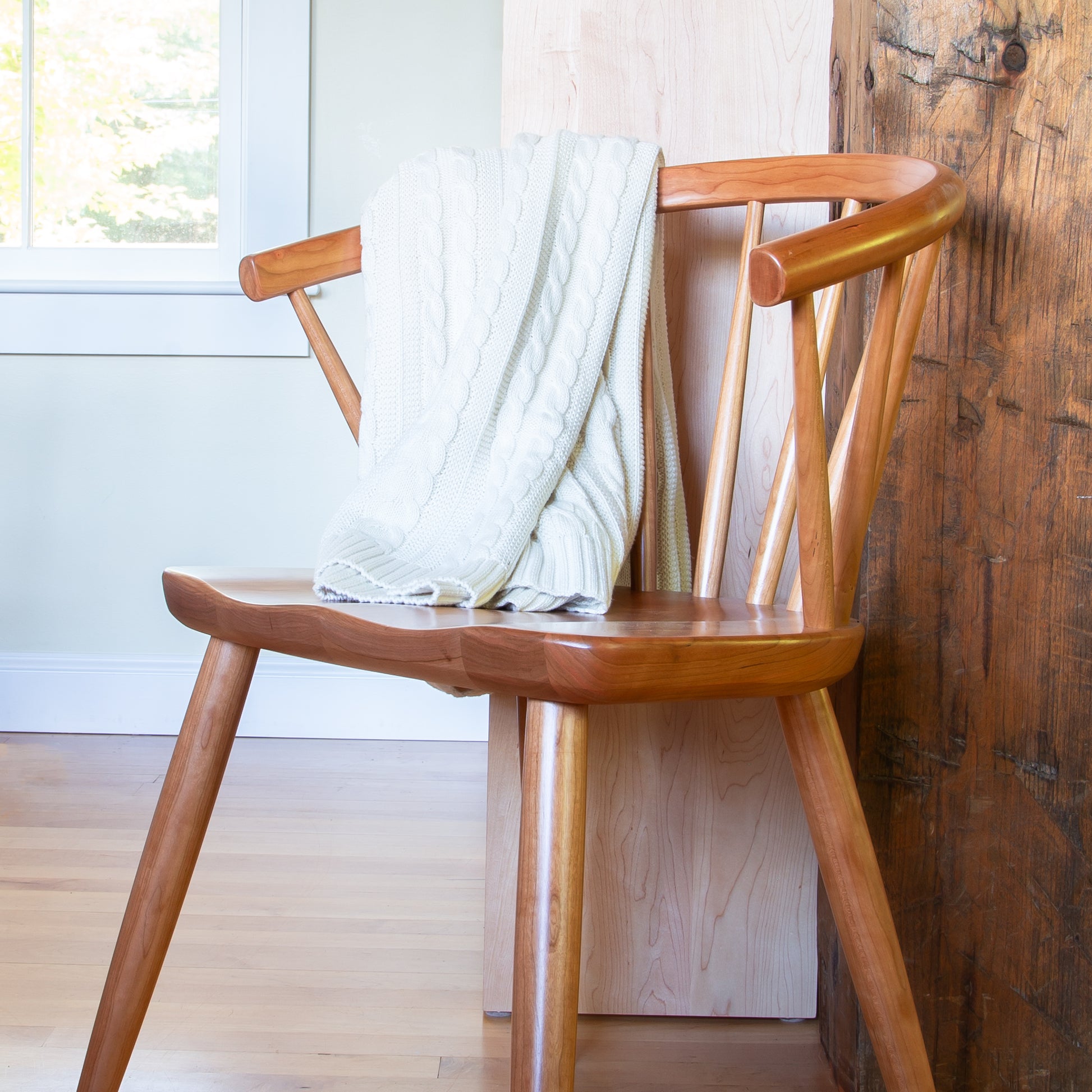 A Dover Curveback Windsor Chair by Vermont Woods Studios, featuring elegantly curved armrests, is placed next to a wooden wall. A white knitted blanket is draped over the backrest. A window in the background allows natural light to illuminate the space, highlighting the wooden floor and light-colored walls.