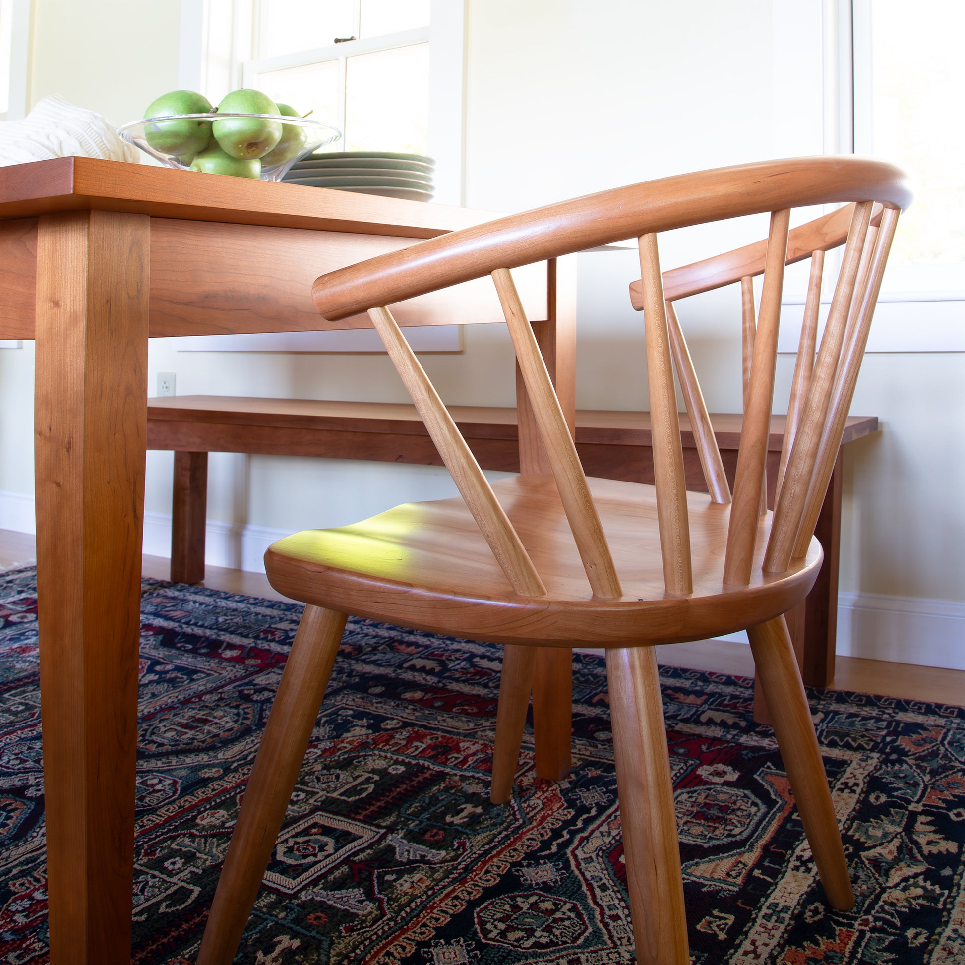 A Dover Curveback Windsor Chair from Vermont Woods Studios with a curved backrest is positioned at a wooden dining table. On the table is a bowl of green apples, a stack of plates, and a window in the background. An intricate patterned rug covers the floor beneath the table and chair.