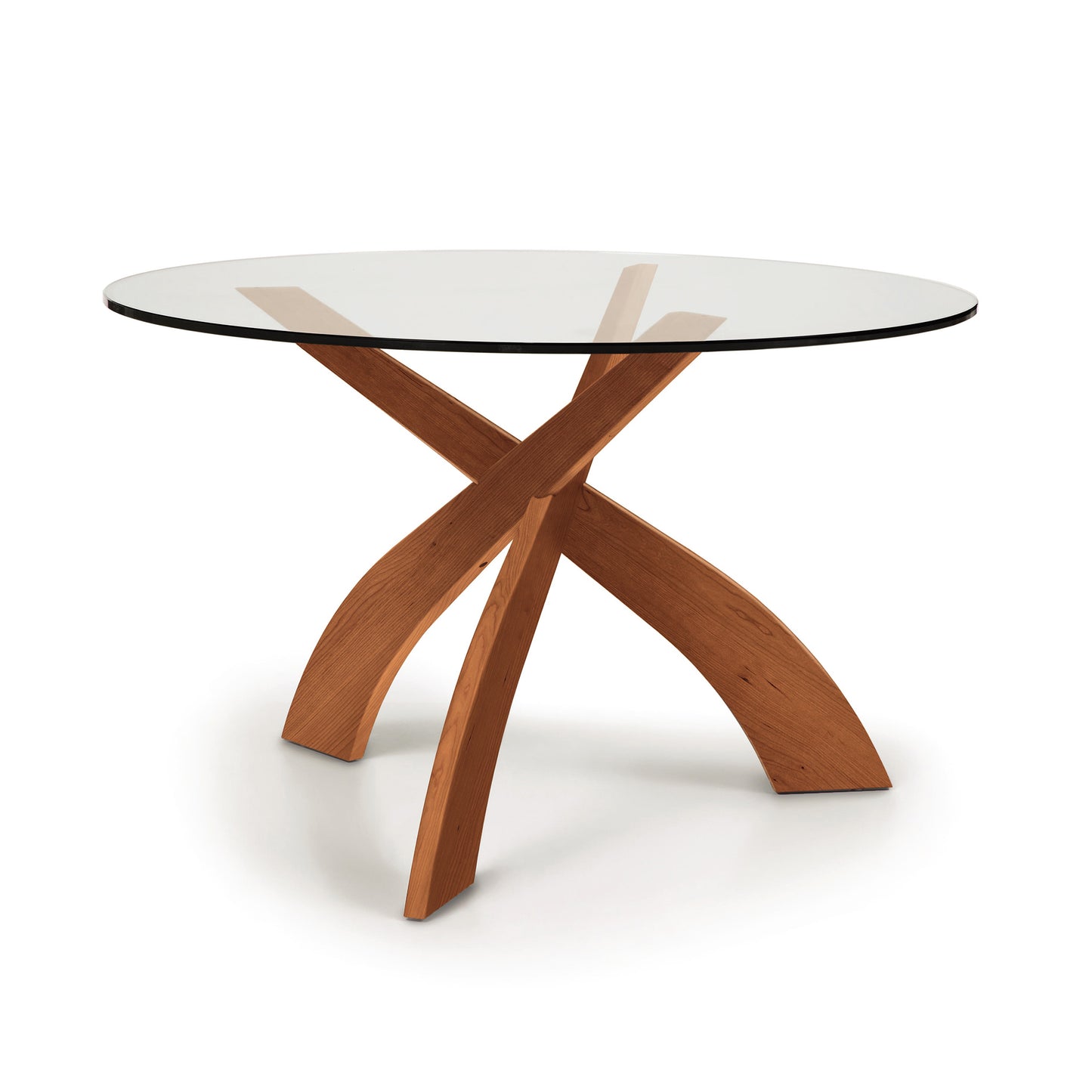 A round tempered glass-top Entwine dining table with a cherry wood, x-shaped base by Copeland Furniture on a white background.