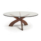 An artistic Copeland Furniture Entwine Round Coffee Table featuring a round glass-top with a wooden cross base on a white background.