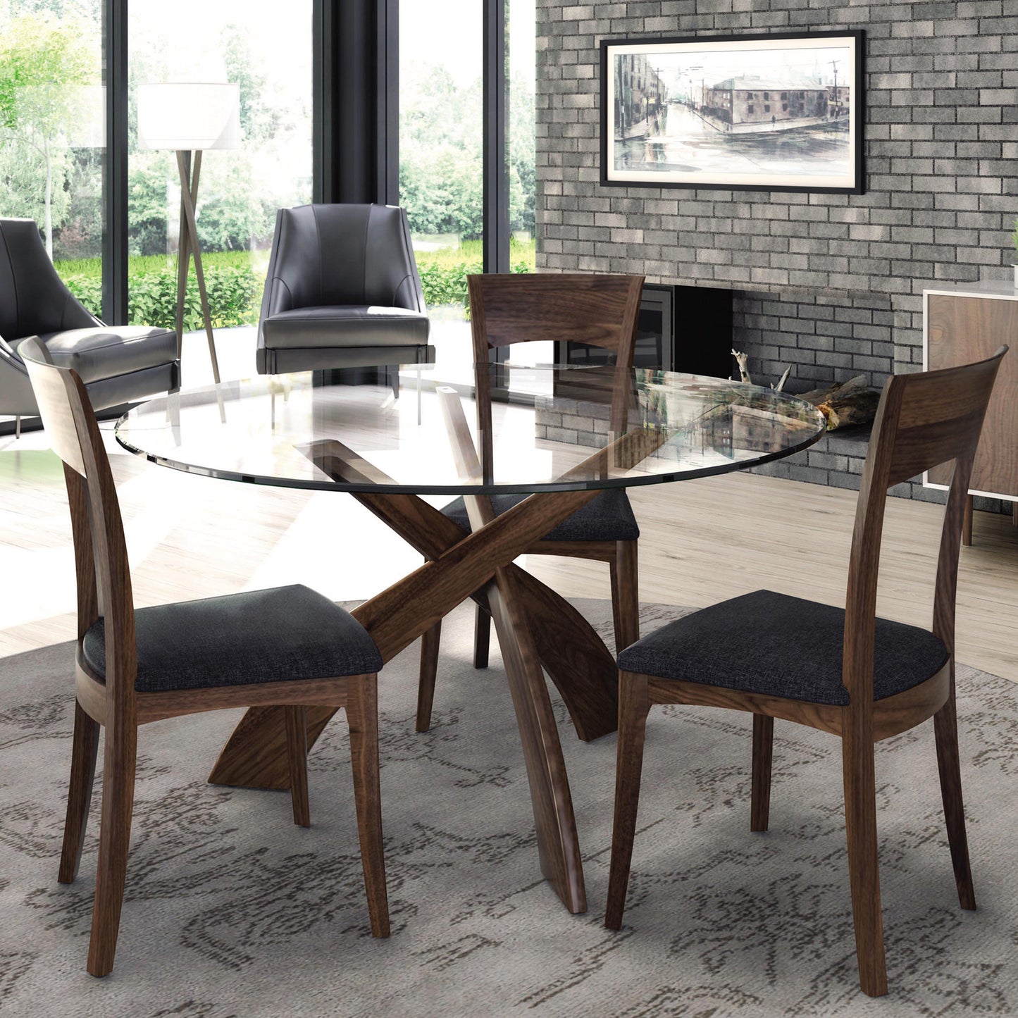 A modern dining room featuring the Entwine Round Glass Top Dining Table by Copeland Furniture, with a tempered glass top and cherry wood base, surrounded by three wooden chairs with dark cushioned seats. The room has a brick wall with a framed picture, two black leather armchairs, a large window, and a floor lamp.