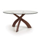 A modern Entwine Round Glass Top Dining Table from Copeland Furniture with a tempered glass top and a sustainably sourced cherry wood cross base isolated on a white background.