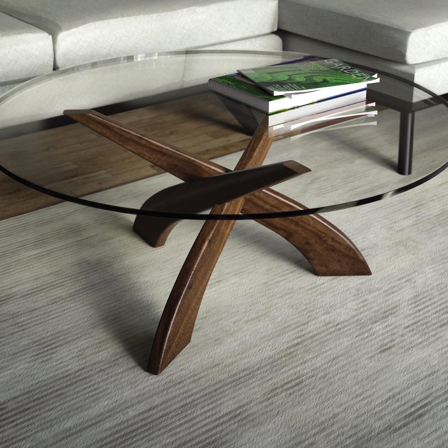 The Entwine Round Coffee Table from Copeland Furniture, featuring a sculptural walnut base and a glass top, is arranged on a light-colored rug in front of a gray sectional sofa. Two books with green and black covers sit on the table, adding a touch of color to the setting.